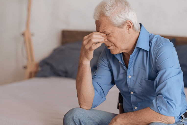 Man with white hair squeezing the bridge of his nose while sitting on a bed. He is suffering from memory loss and headaches due to sleep apnea