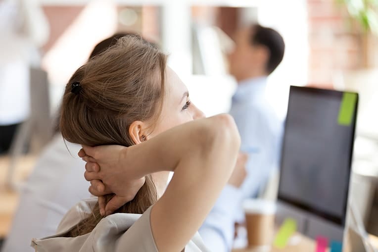 Woman with her hands on her back stretches while working at her computer. Maintaining good posture is just one habit you should break in order to help your TMJ and TMD symptoms. 